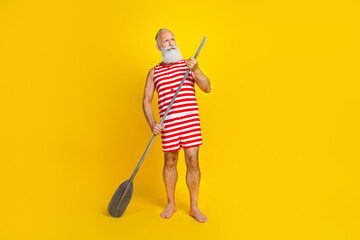 Full length photo of tourist old man kayaking looking beautiful natures surfing sap board with oar isolated on yellow color background