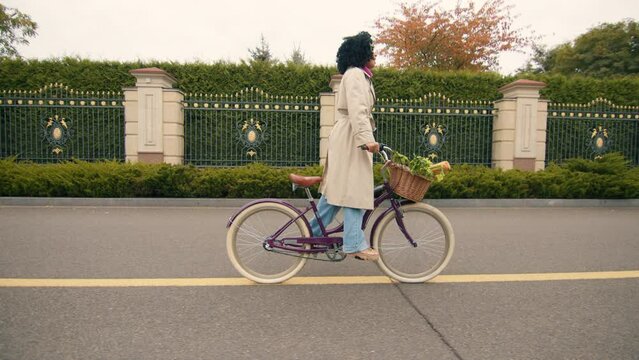 Happy pretty African woman in autumn trendy outfit on a bike ride along road, passing by beautiful metal fence with natural privacy fence, yellow trees. High quality 4k footage