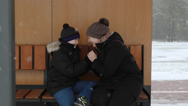 Mother warms son hands on bench on frosty winter day. Happy family smiles and touches each other foreheads and noses. Relaxing together and taking care on snowy day in the city.
