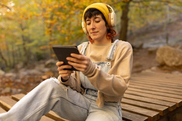 Woman with mobile phone and headphones in forest wearing yellow hat, listening to music, taking photos and video call
