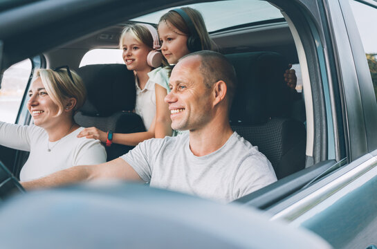 Happy young couple with two daughters inside the modern car with panoramic roof during auto trop. They are smiling, laughing during road trip. Family values, traveling concepts..