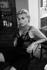 Fototapeta na wymiar A beautiful slender young mysterious blonde sits at a table in a summer cafe. Black and white fashion photography.