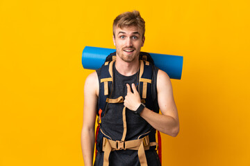 Young mountaineer man with a big backpack isolated on yellow background with surprise facial...