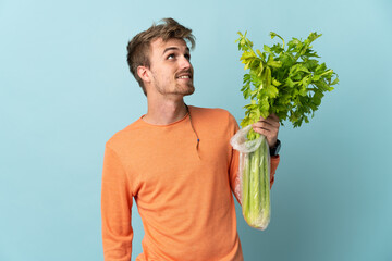Young blonde man holding a celery isolated on blue background looking up while smiling