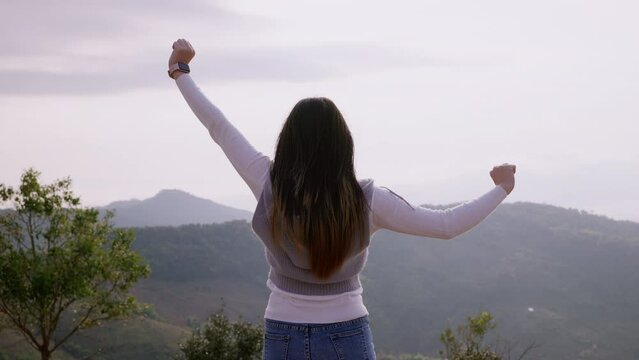 Back view of hipster millennial young asian woman on top of mountain summit at sunset raises arms into air happy and drunk on life youth and happiness.