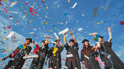 Graduates throw colorful confetti against a blue sky.