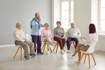 Senior people in group therapy. Happy retired old men and women sitting on chairs in a light office or retirement house and talking to a young friendly woman therapist with a clipboard - obrazy, fototapety, plakaty