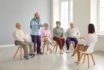 Zelfklevend Fotobehang Senior people in group therapy. Happy retired old men and women sitting on chairs in a light office or retirement house and talking to a young friendly woman therapist with a clipboard © Studio Romantic