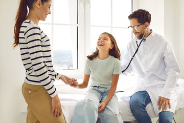 Male smiling family doctor with stethoscope examining child's lungs, breathing and heartbeat sitting on the couch with her mother in medical clinic. Pediatrician checking girl patient in exam room.