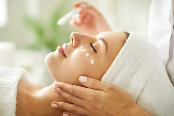 Close up beautician's hands applying anti-aging facial cream on woman client face to prevent...