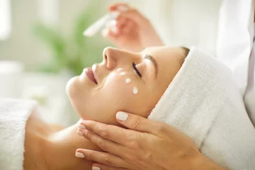 Badezimmer Foto Rückwand Spa Close up beautician's hands applying anti-aging facial cream on woman client face to prevent wrinkles in spa salon. Skin care, cosmetic procedures for facial care and beauty treatment concept.
