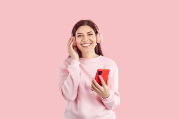 Young positive Caucasian woman melomaniac in wireless headphones enjoys listening to music and smiling broadly holding mobile phone dressed in casual style stands on studio pink background