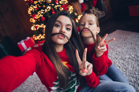 Photo of cute shiny small sisters wear red jumpers recording video vlog showing v-sign fooling indoors new year north pole house