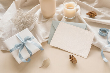 Holiday background with blank paper card, envelope, gift box on linen fabric, gift, fir cones, ribbon, candle.