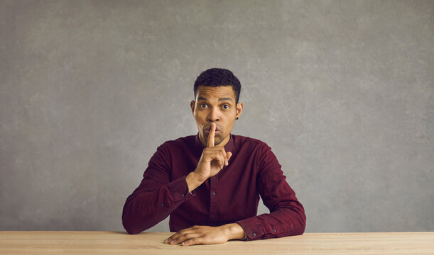 Don't tell anyone. Studio shot of handsome young black man sitting at desk, looking at camera and doing finger on lips shush gesture asking you to keep your mouth shut, be silent and keep his secret