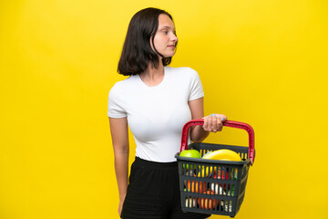 Young woman holding a shopping basket full of food isolated on yellow background looking to the side
