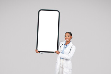 Positive millennial black lady doctor therapist in white coat hold big mobile phone