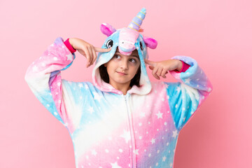 Little caucasian woman wearing a unicorn pajama isolated on pink background having doubts and...