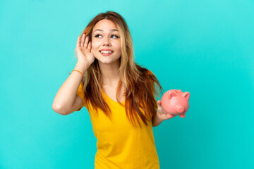 Teenager blonde girl holding a piggybank over isolated blue background listening to something by...