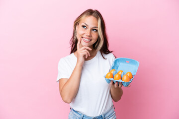 Young Russian woman holding eggs isolated on pink background thinking an idea while looking up