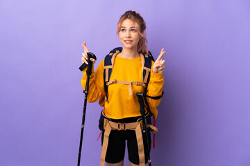Teenager girl with backpack and trekking poles over isolated purple background with fingers crossing and wishing the best