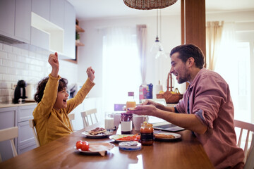 Happy father and little son having breakfast together at home kitchen - obrazy, fototapety, plakaty