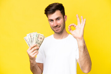 Young caucasian man taking a lot of money isolated on yellow background showing ok sign with fingers
