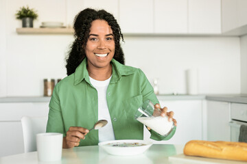 african woman pouring milk into bowl with cereals in kitchen - obrazy, fototapety, plakaty