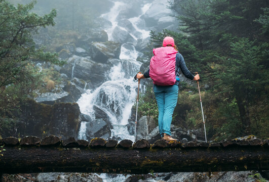 Young woman with backpack and trekking poles enjoying power mountain river waterfall on wooden bridge during Makalu Barun National Park trek in Nepal. Mountain hiking and active people concept image