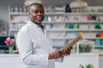 Portrait of a happy African-American senior pharmacy owner standing in a drug store with a clipboard in his hands and smiling at the camera. - obrazy, fototapety, plakaty
