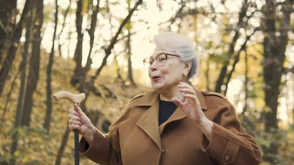 Active mature woman holding walking cane in hands and dancing, actively moving to the music rhythm, pleasure pastime in autumn park, good mood, positivity and energy charge