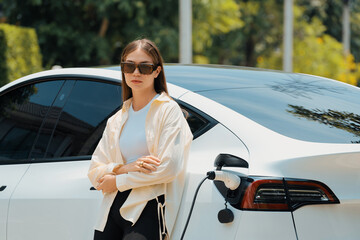 Young woman recharge her EV electric vehicle at green city park parking lot. Urban sustainability...