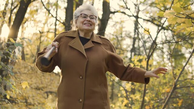 Happy mature lady with gray hair in warm coat actively dancing outdoor, holding walking cane in hands, celebrating birthday, anniversary or just taking part in street performance