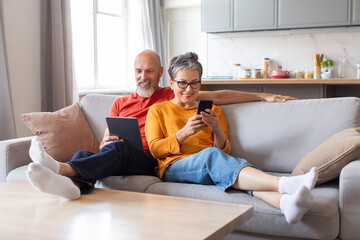 Smiling old couple resting on sofa with digital tablet and mobile phone