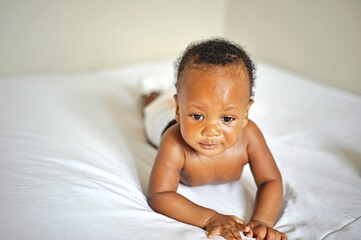 Childcare Concept. Portrait of cute little African  baby wearing bodysuit lying on white bedsheets...