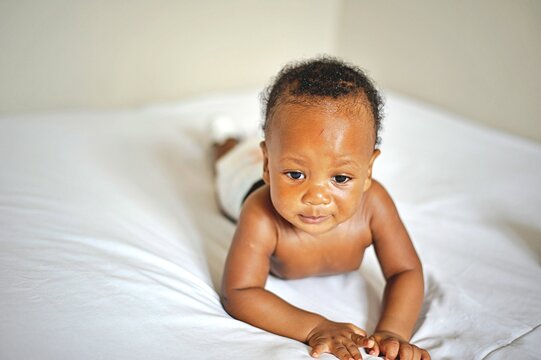 Childcare Concept. Portrait of cute little African  baby wearing bodysuit lying on white bedsheets at home. Black infant child crawling on bed in the bedroom. Selective focus, free copy space