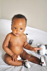 Childcare Concept. Portrait of cute little African  baby wearing bodysuit lying on white bedsheets at home. Black infant child crawling on bed in the bedroom. Selective focus, free copy space