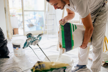 A handyman is adding plaster in a bucket and preparing it for skim coating. - obrazy, fototapety, plakaty