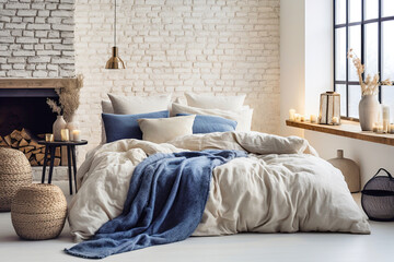 Bed with blue pillows and coverlet near fireplace against white brick wall. Loft, scandinavian interior design of modern bedroom. - obrazy, fototapety, plakaty
