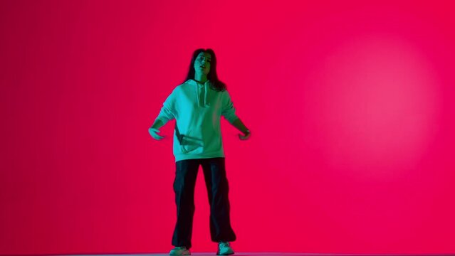 Attractive woman in pants and hoodie dancing dynamic jazz funk choreography, isolated on red background neon light.