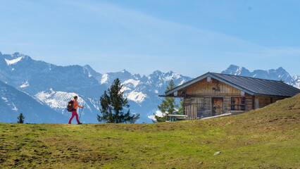 Auf dem Staffel Berg in den Bayerischen Voralpen läuft eine Wanderin zu einer Hütte mit schneebedeckten Bergen im Hintergrund.
