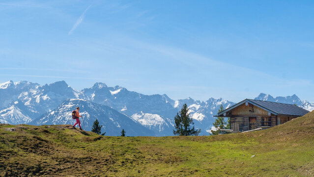 Auf dem Staffel Berg in den Bayerischen Voralpen l&auml;uft eine Wanderin zu einer H&uuml;tte mit schneebedeckten Bergen im Hintergrund.