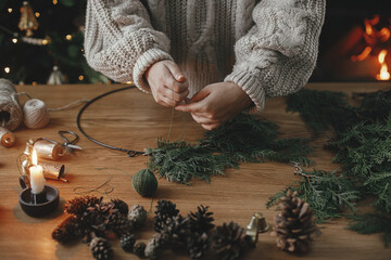 Hands in cozy sweater making Christmas rustic wreath with fir branches, pine cones, candle, twine,...