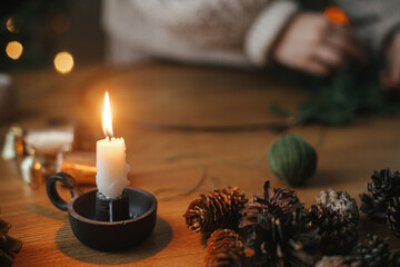 Candle, pine cones, bells, twinq on wooden table in evening festive room. Making Christmas rustic...