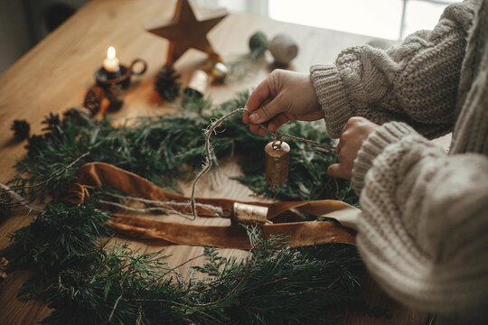 Hands in cozy sweater making Christmas rustic wreath with fir branches, ribbon, pine cones, bells on wooden table, close up. Winter holiday preparations, atmospheric time