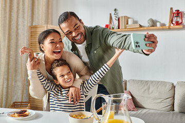 modern cheerful african american family taking selfies at breakfast table and smiling at camera