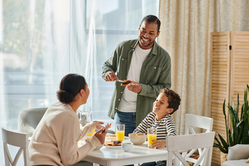 cheerful african american man spreading jam on toast and looking at his wife and son at breakfast - obrazy, fototapety, plakaty