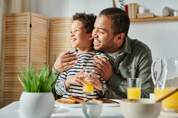 happy african american father and son in casual homewear hugging at breakfast table looking away