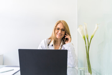 Smiling female doctor talking on the phone to a patient while looking at camera in medical office.