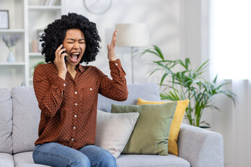 Frustrated and angry woman talking on the phone, African American woman shouting and angry at the interlocutor, sitting on the sofa in the living room of the house alone - obrazy, fototapety, plakaty